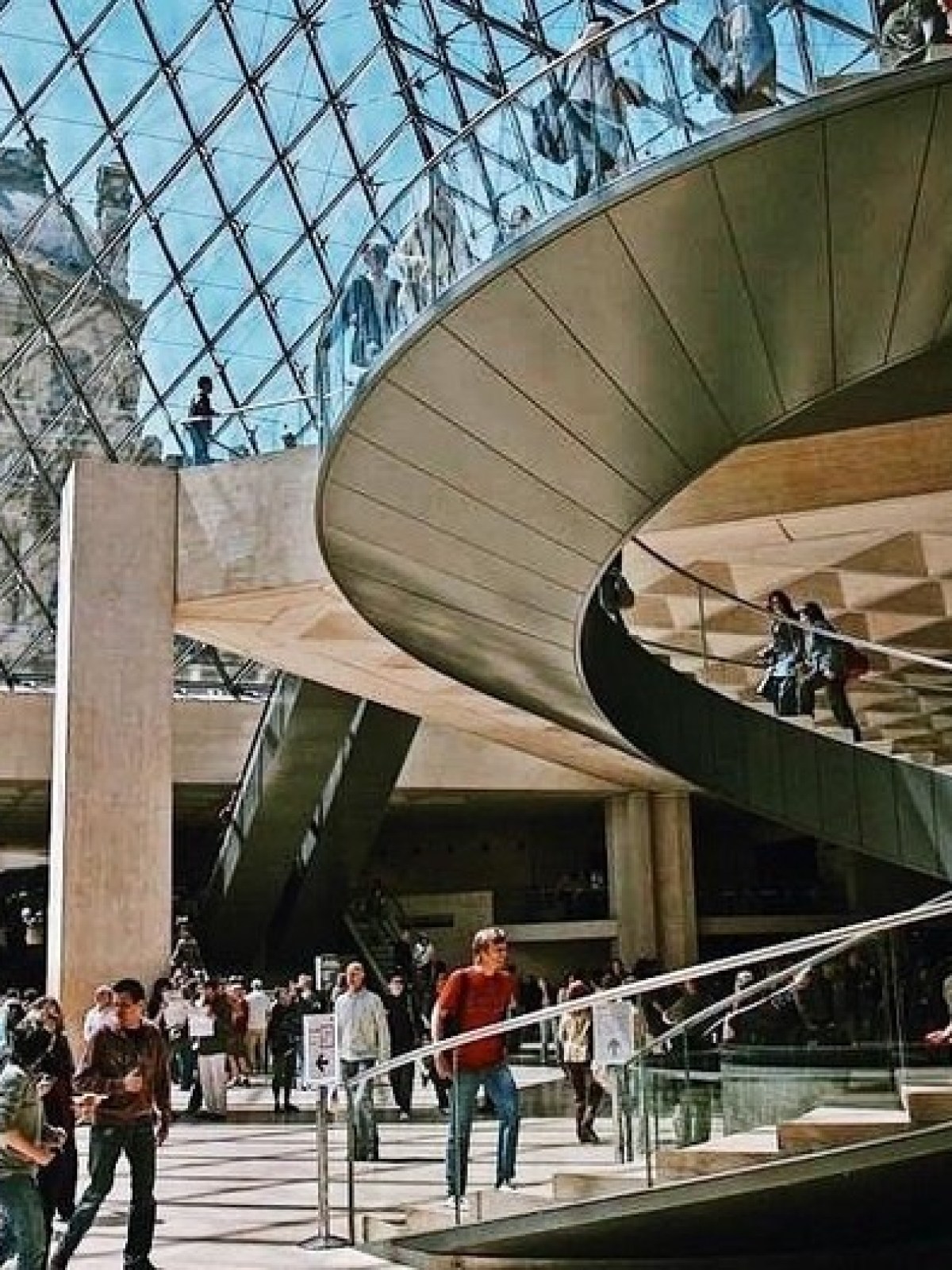 People walking on a spiral staircase under a glass pyramid with a classical building in the background.