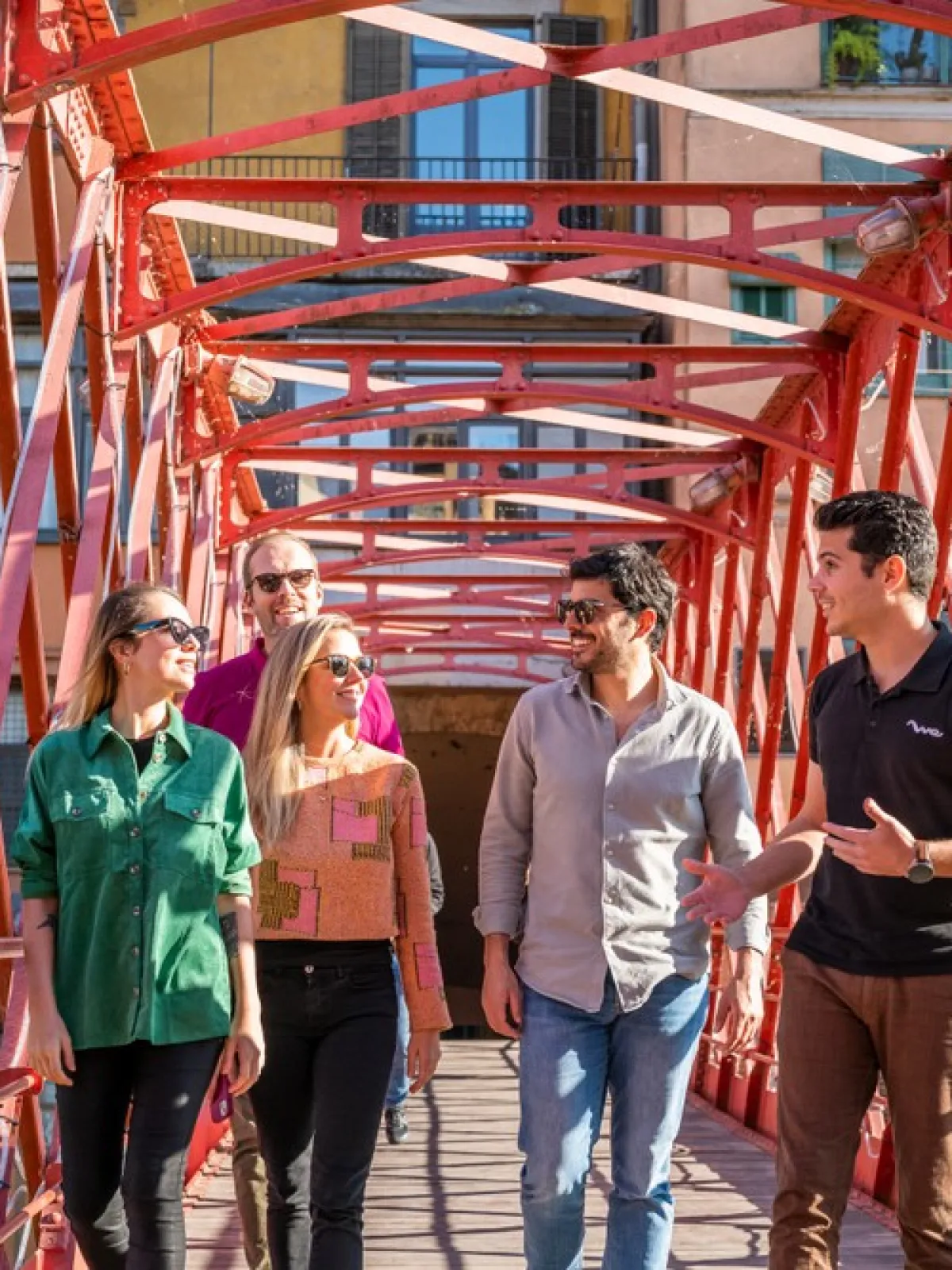 Five people walking on a red metal bridge, surrounded by buildings.