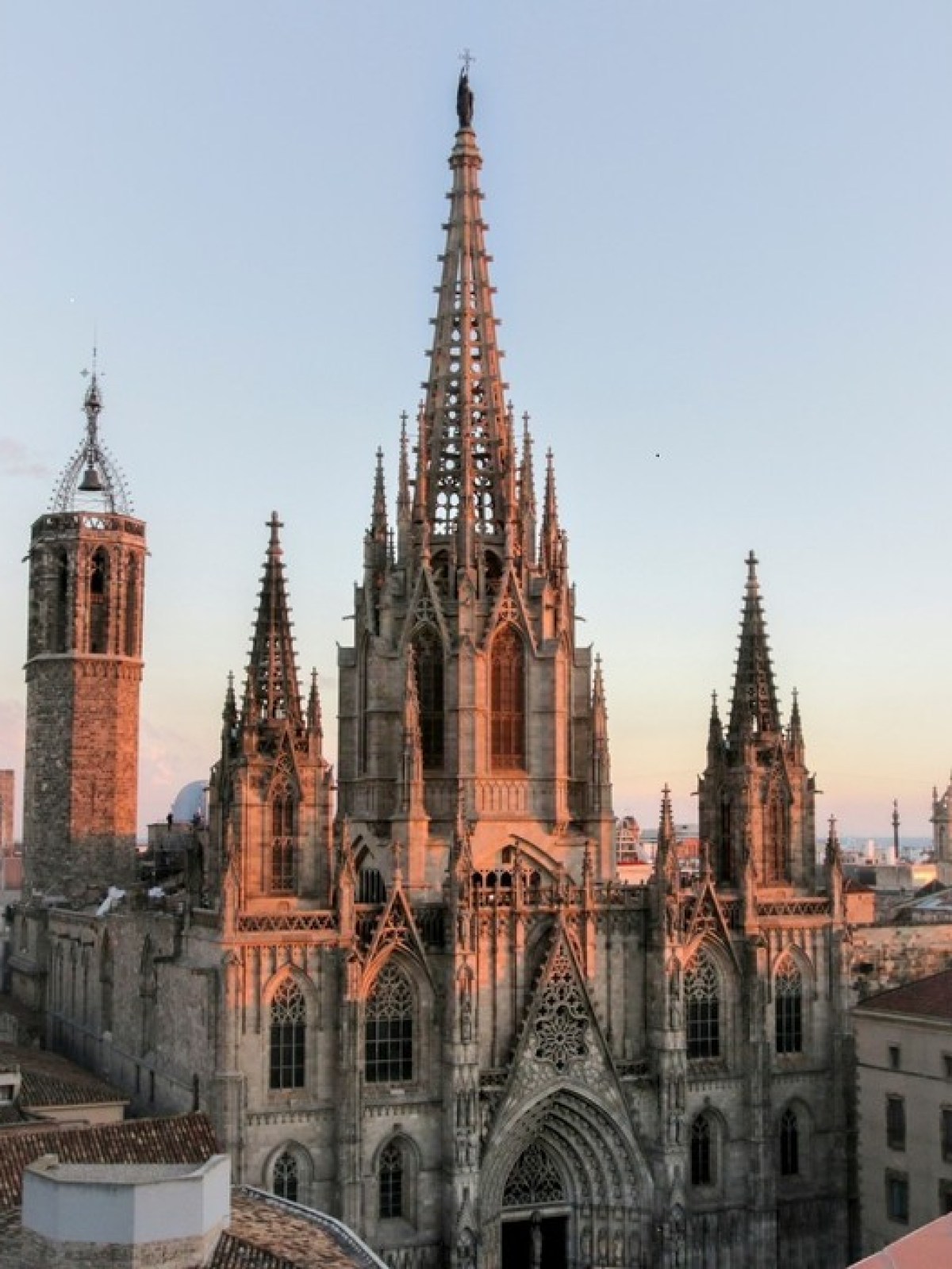 Gothic cathedral with towers and spires at sunset, surrounded by city rooftops.