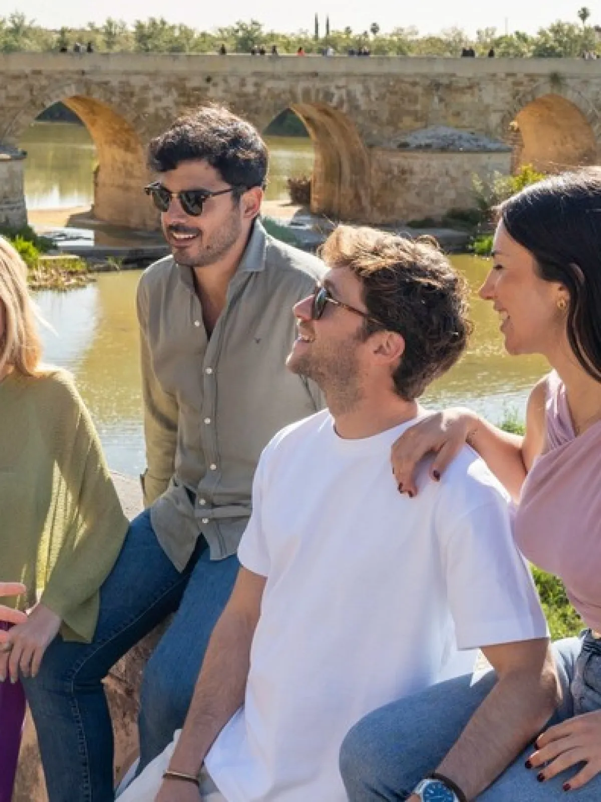Five people talking and smiling by an ancient stone bridge over a river.