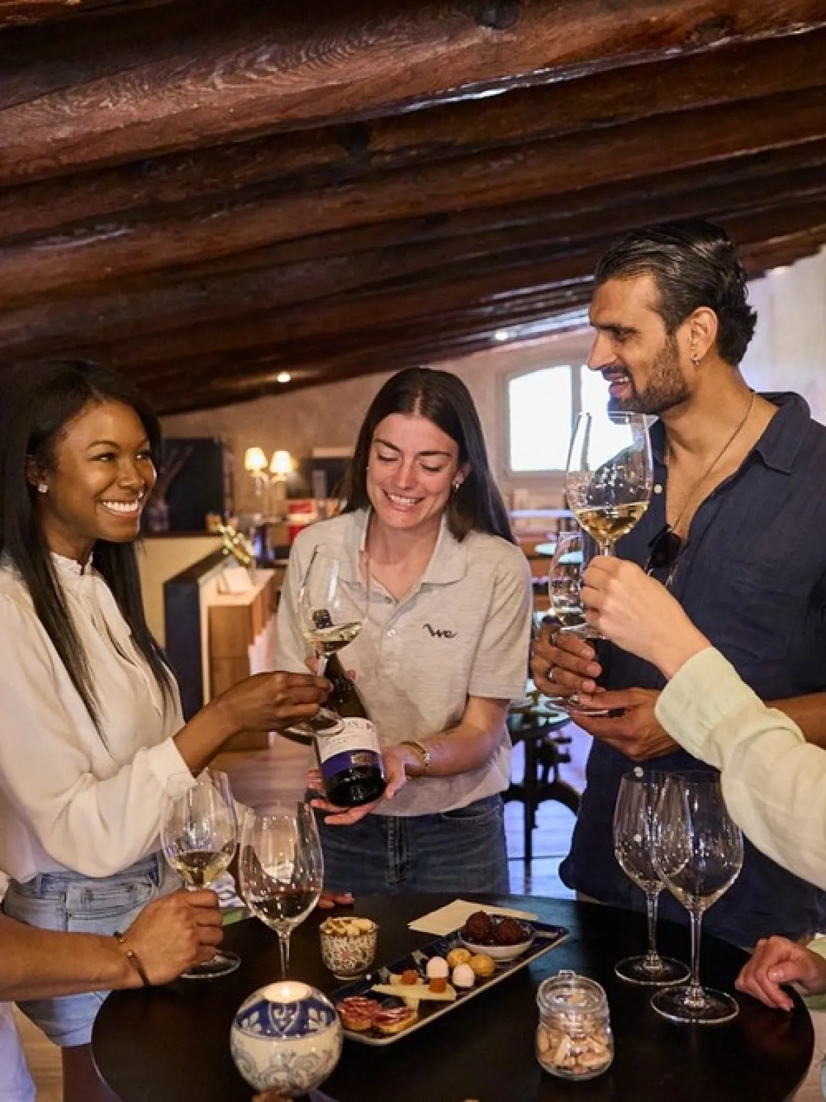 Five people smiling and toasting with wine glasses in a cozy room with wooden beams.