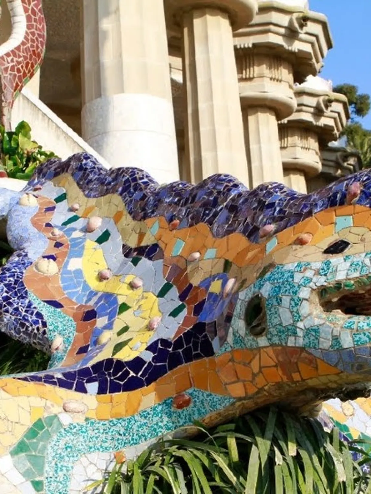 Colorful mosaic lizard sculpture on steps with greenery, surrounded by architectural columns and trees.