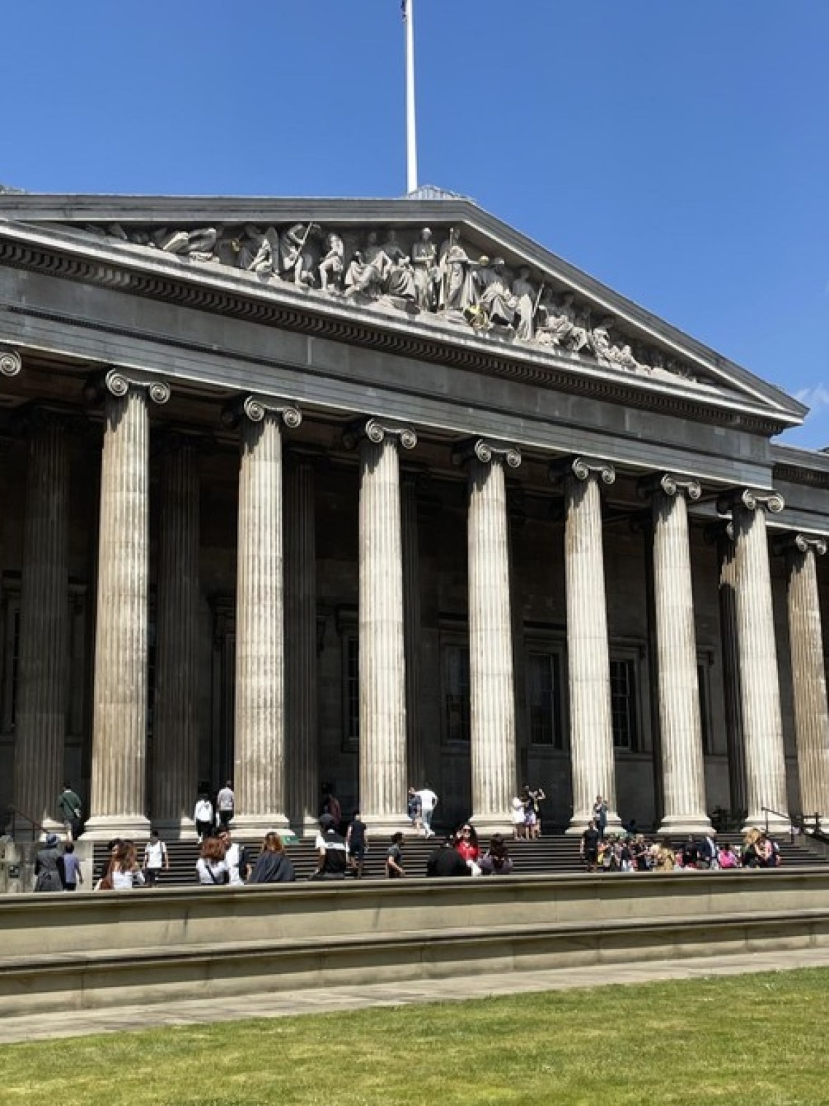 Neoclassical building with columns and people sitting outside on a sunny day.