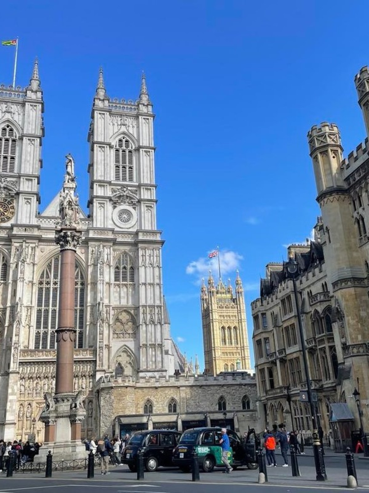 Historic gothic buildings with towers under a clear blue sky, people and cars in the foreground.