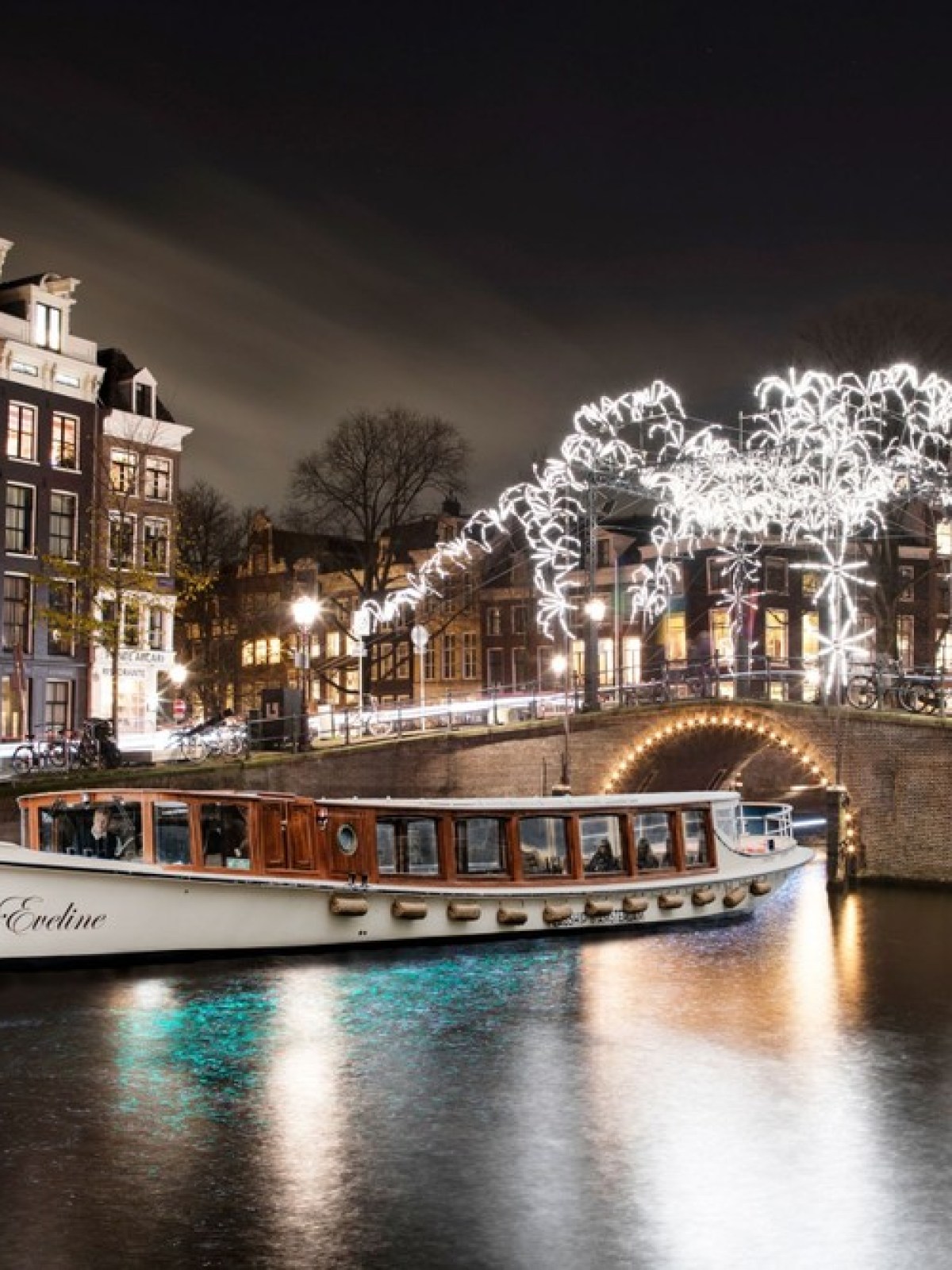 Boat on illuminated canal at night with arched bridge and festive lights in cityscape.