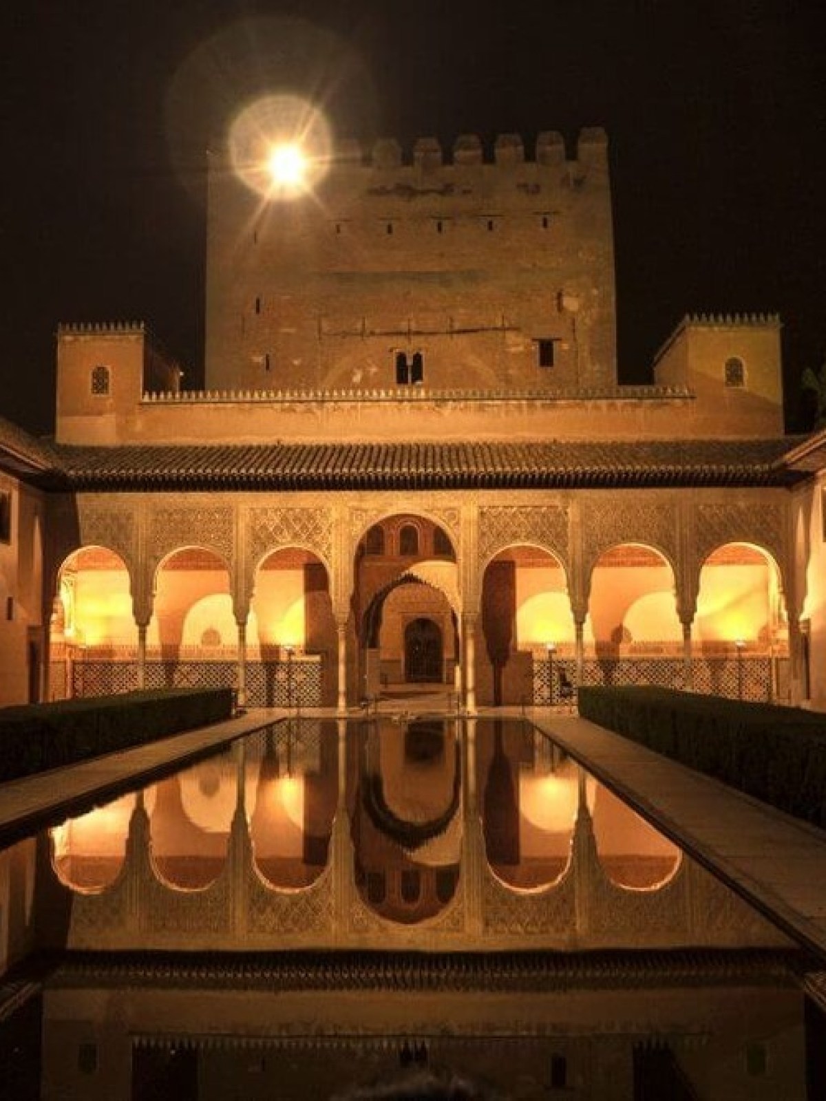 Illuminated courtyard with arches and a reflecting pool at night.