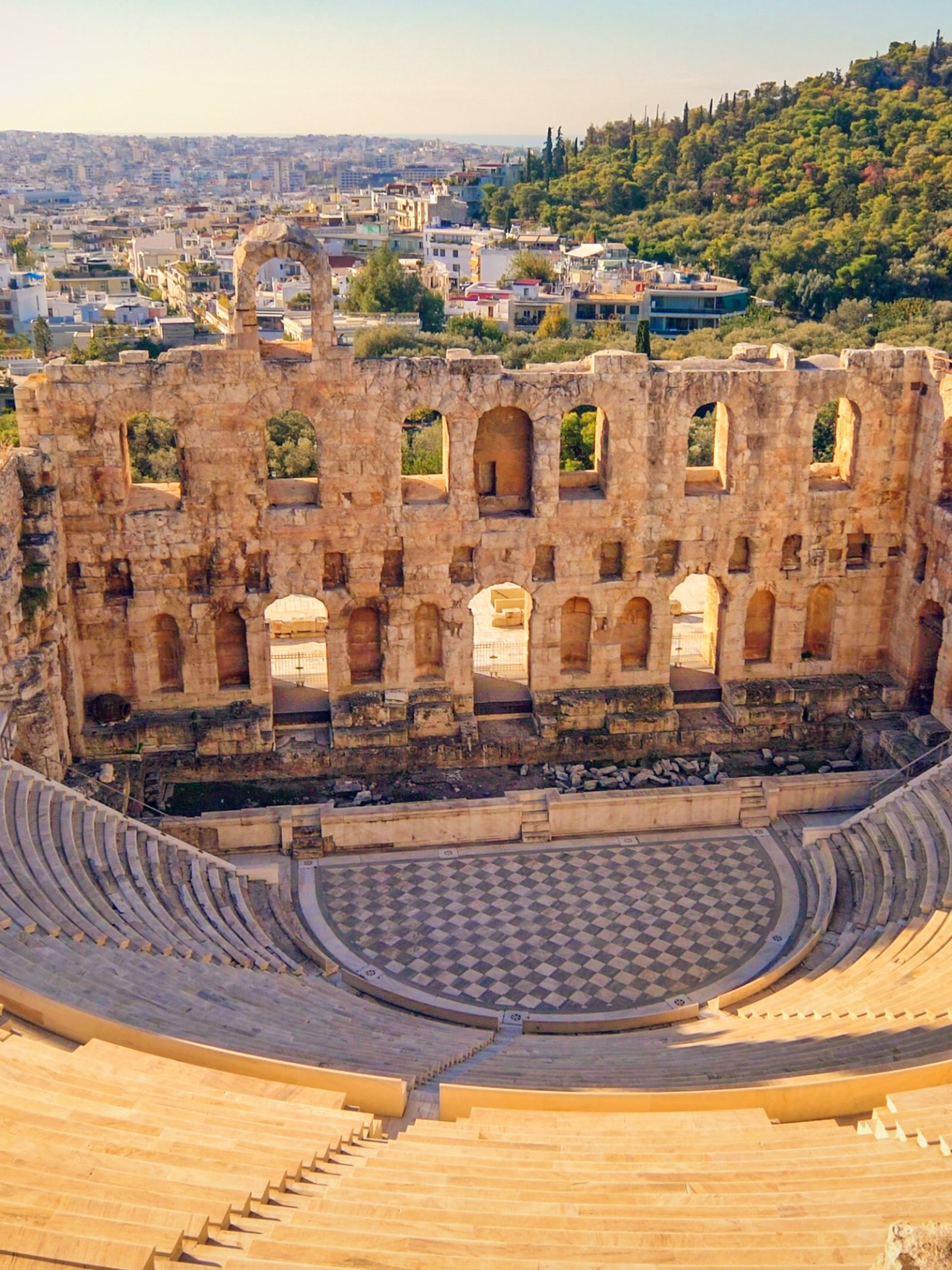 Ancient amphitheater with stone seating and arched ruins, overlooking a cityscape and hillside.
