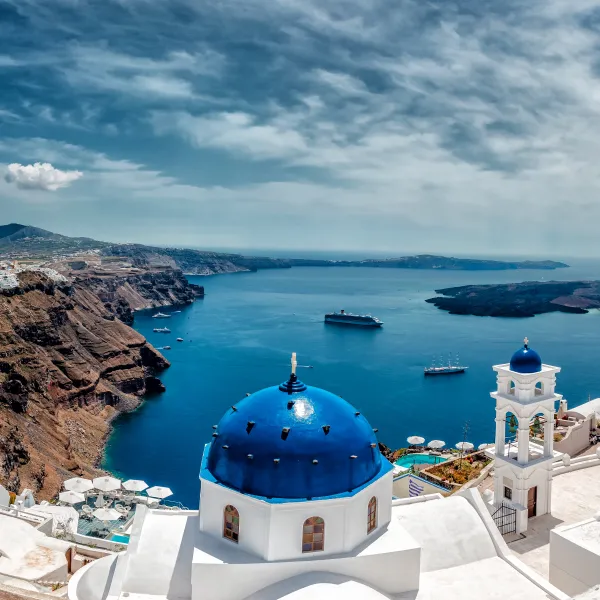 Panoramic view of Santorini with white buildings, blue domes, cliffs, and sea under a cloudy sky.