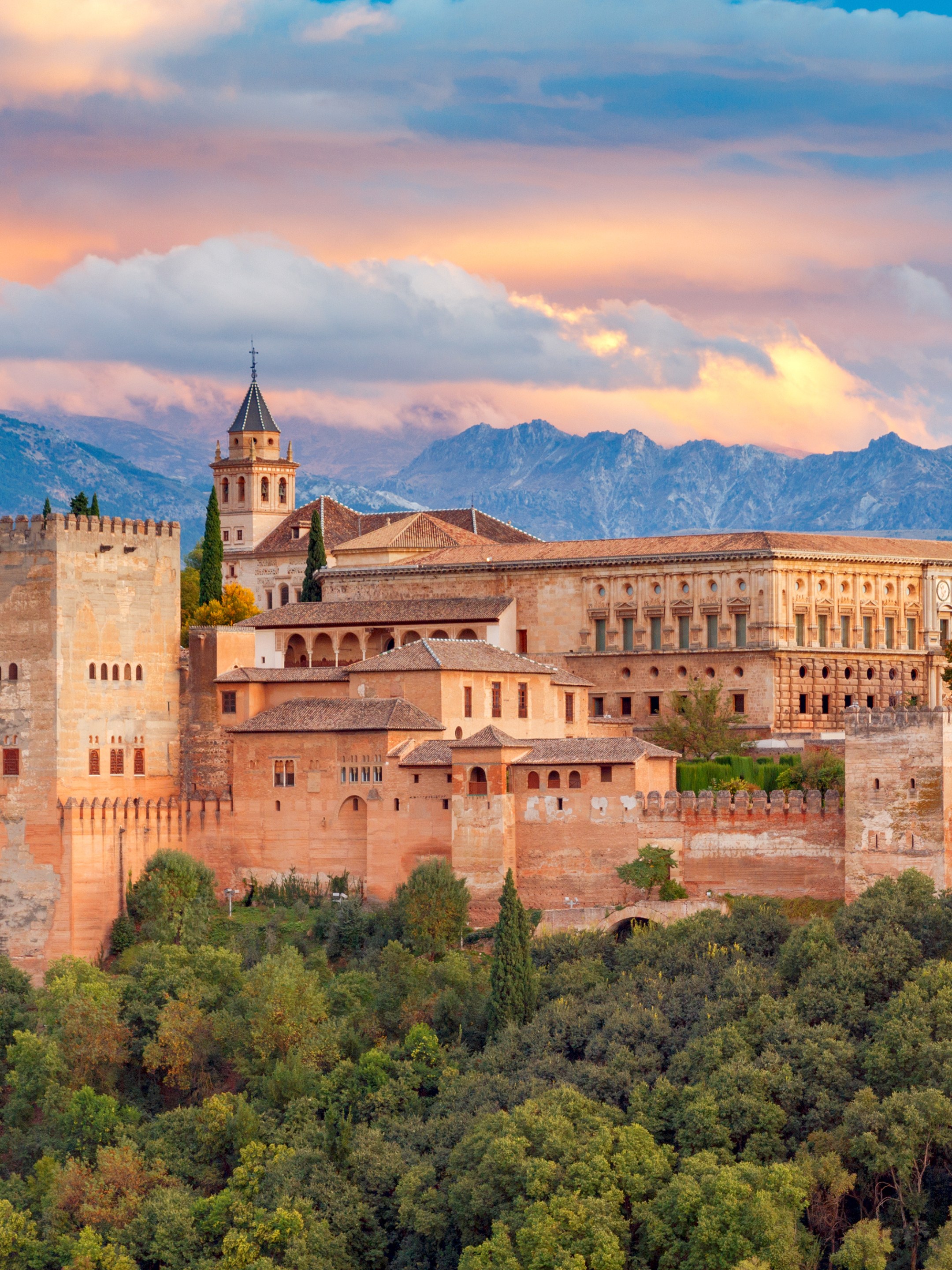 Historic castle with towers and trees at sunset with mountains in the background.