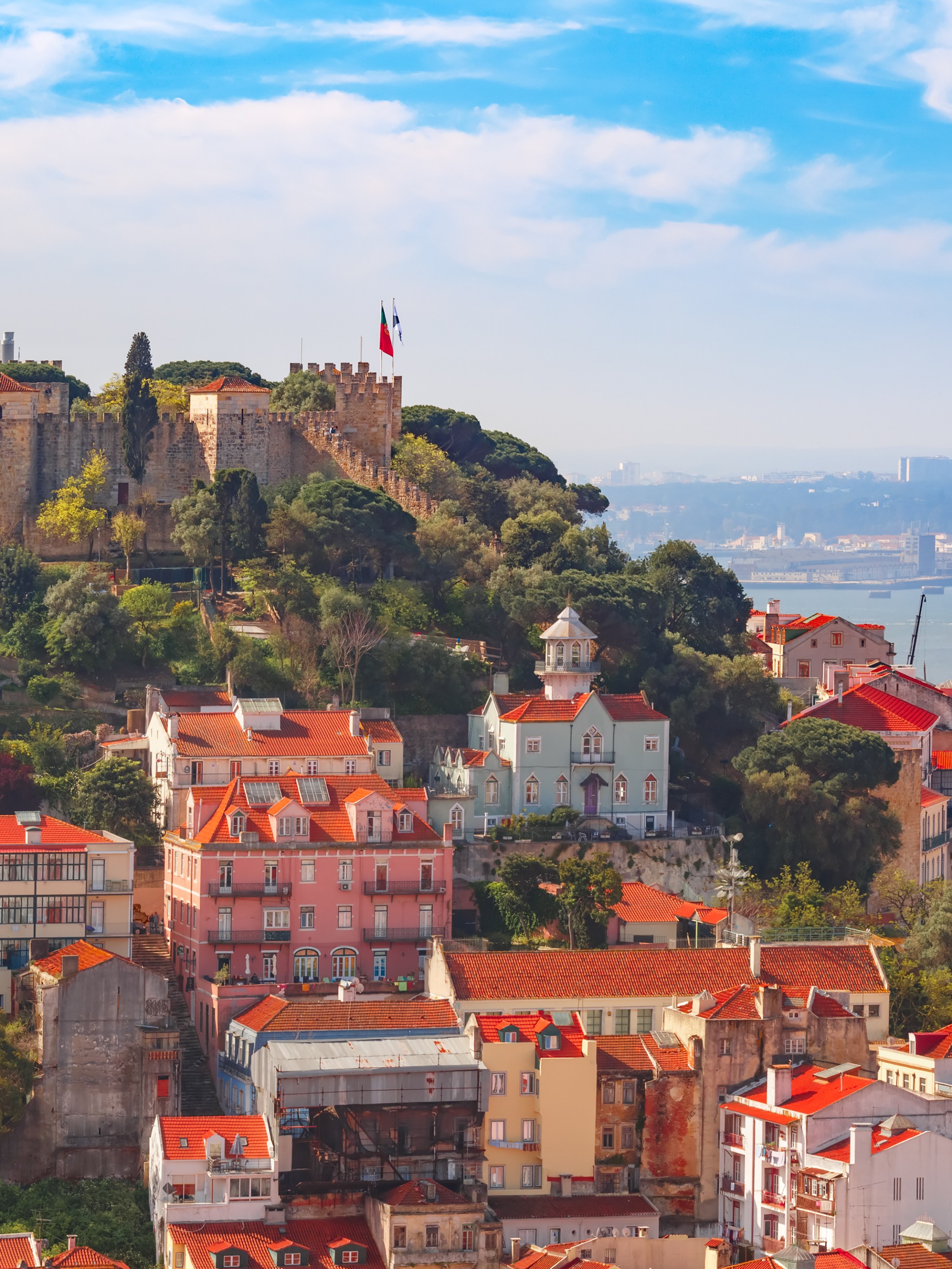 Hilltop castle and colorful houses with red roofs overlooking a river and cityscape.