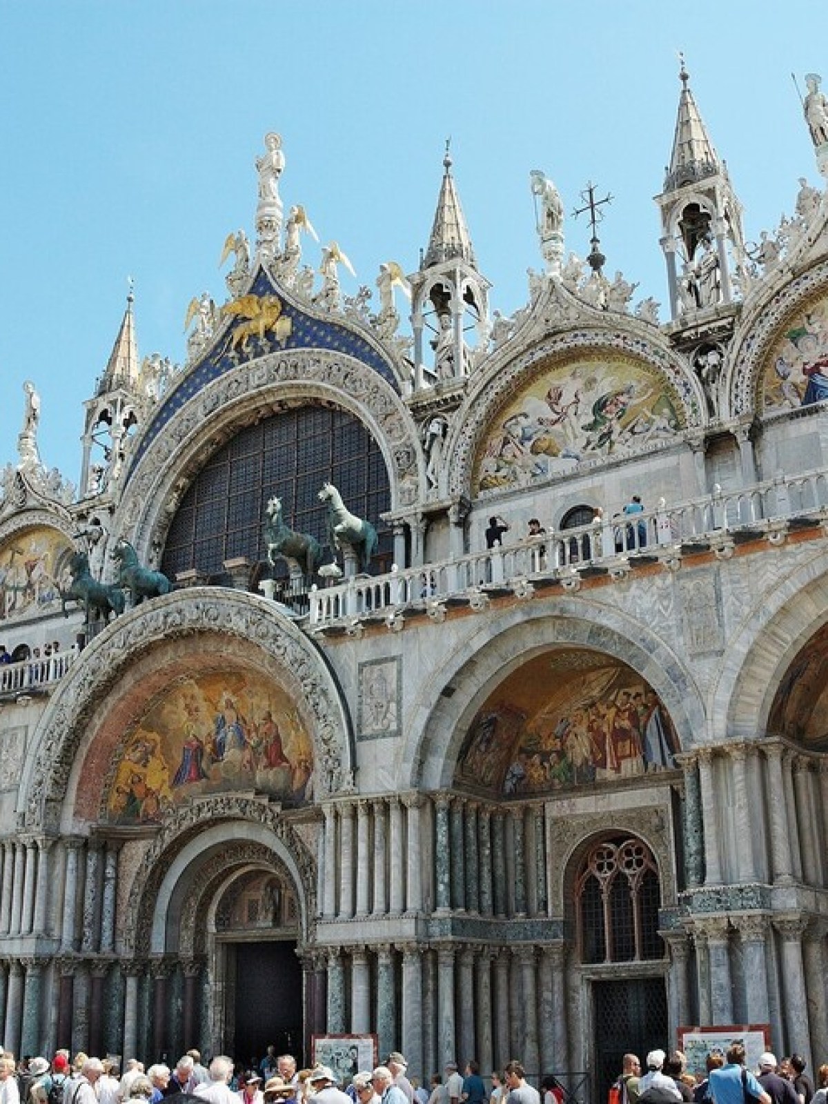 Crowds gather outside Saint Mark's Basilica with ornate facade and sculptures under clear sky.