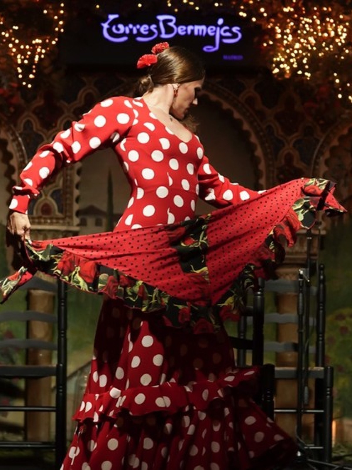 Flamenco dancer in red polka dot dress performing on stage with festive lights.