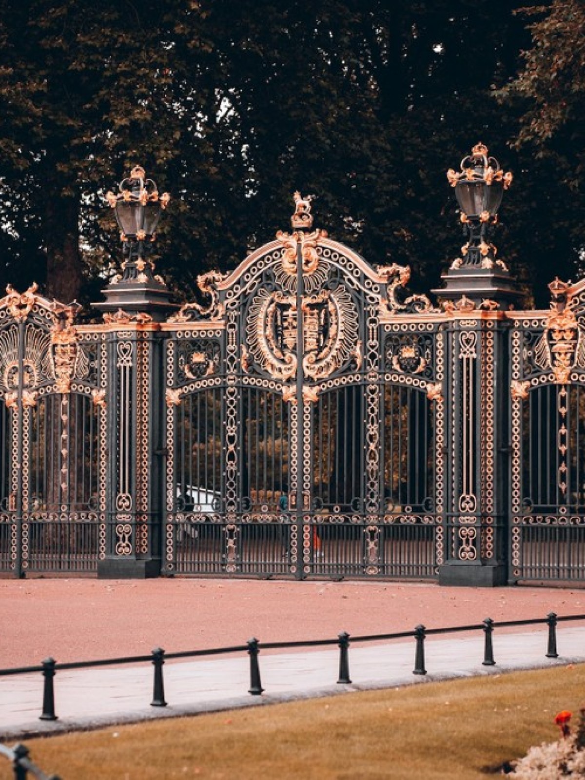 Ornate black and gold gate with intricate designs; trees and flower bed in the background.