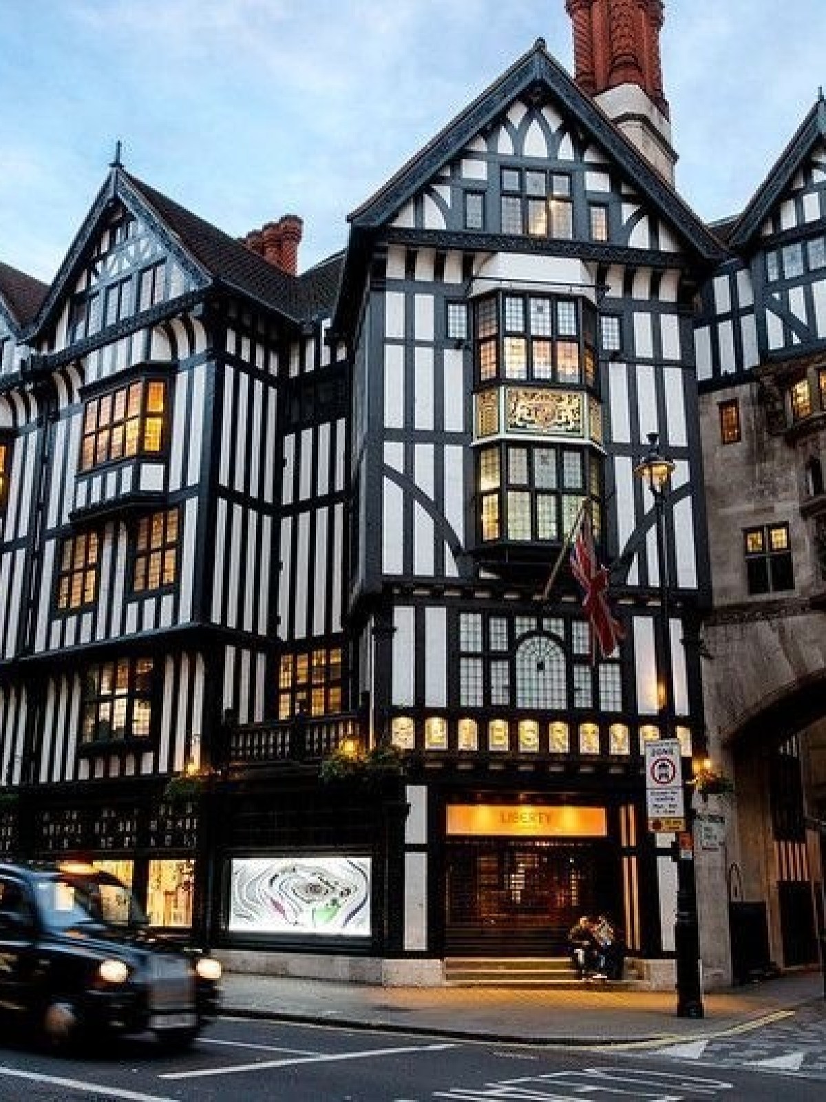 Tudor-style building with shops and a taxi driving by on a busy street at dusk.