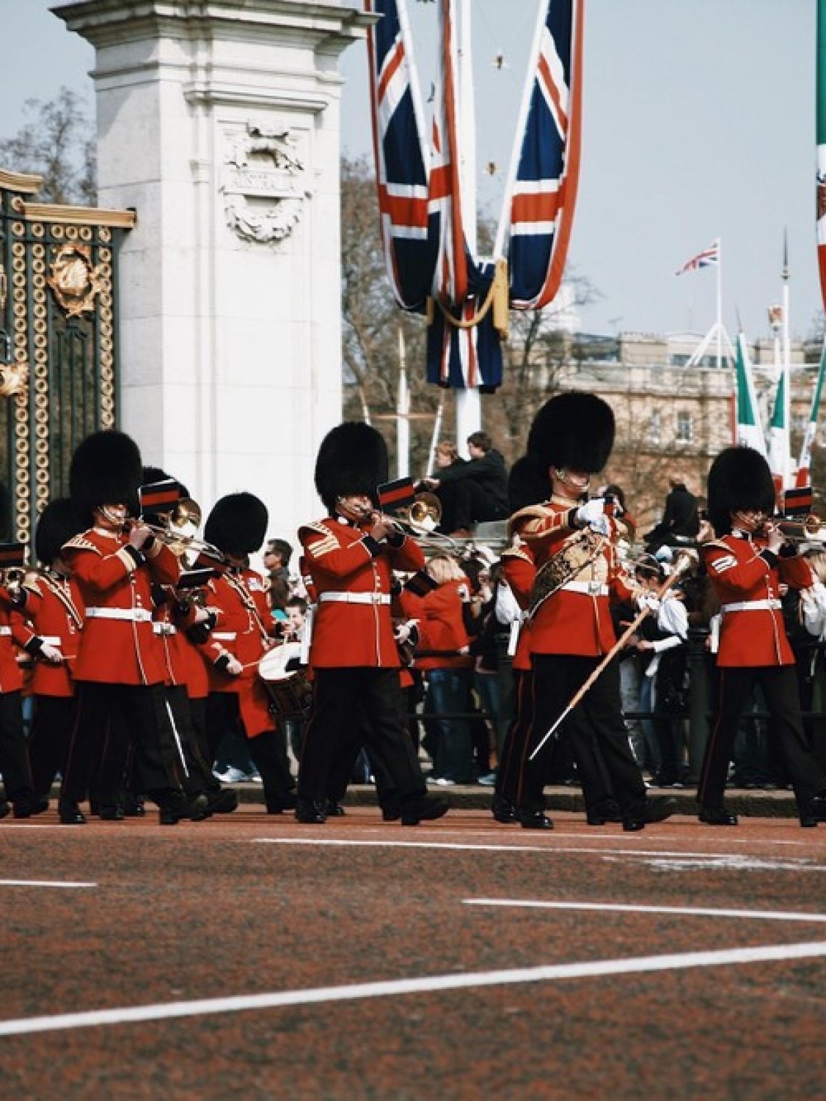 Marching band in red uniforms with bearskin hats in front of ornate gates and flags.