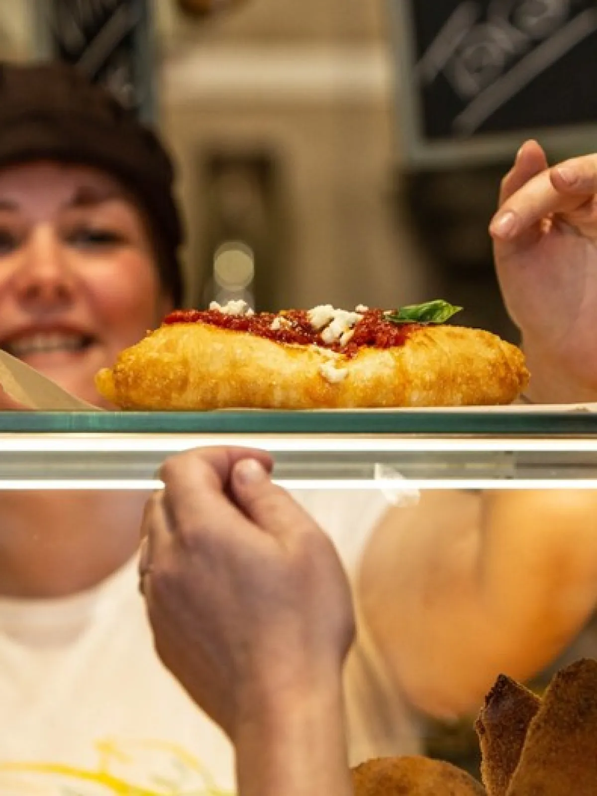 Person handing a fried pizza topped with tomato and basil over a counter in a food shop.