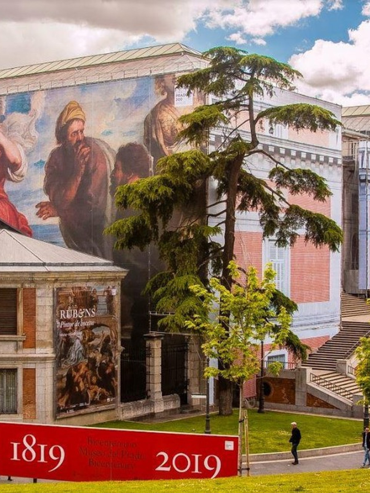 Large building with classical artwork murals and a red sign reading '1819 - 2019' in a landscaped outdoor area.