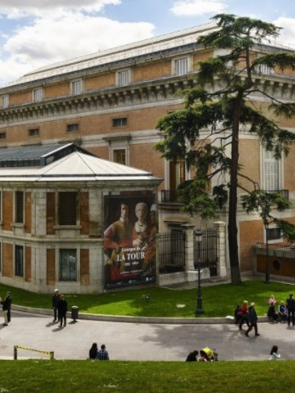 Historic museum building with people on pathways and stairs, surrounded by grass and trees, under a partly cloudy sky.