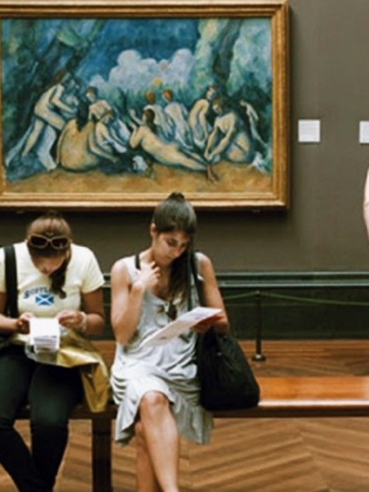 People viewing paintings in a museum with art on the walls and a wooden bench.