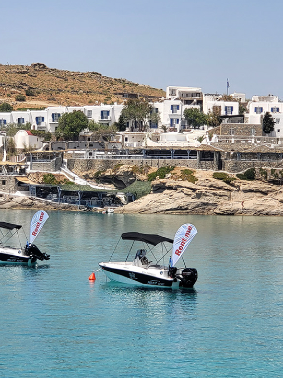 Two small motorboats float in clear blue water near white buildings on a rocky coast.