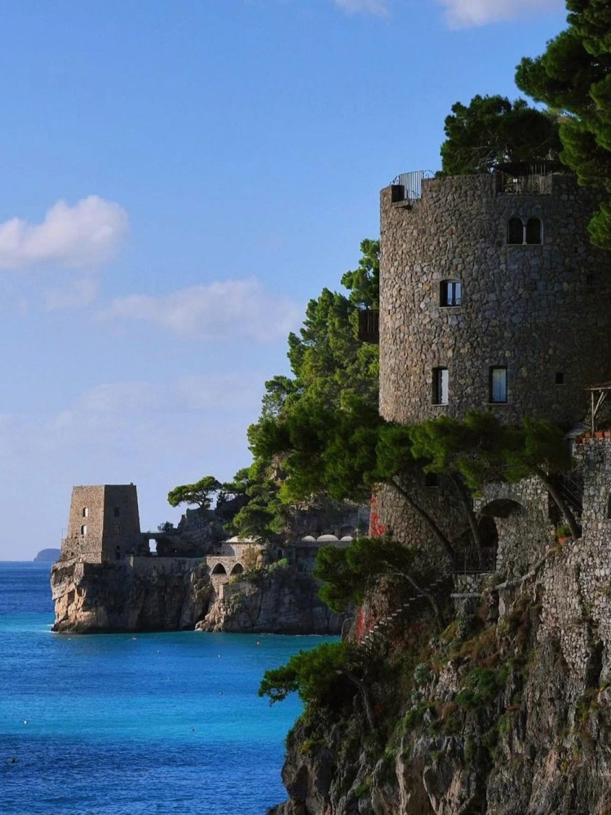 Stone towers and green trees on a cliffside above turquoise sea under blue sky.