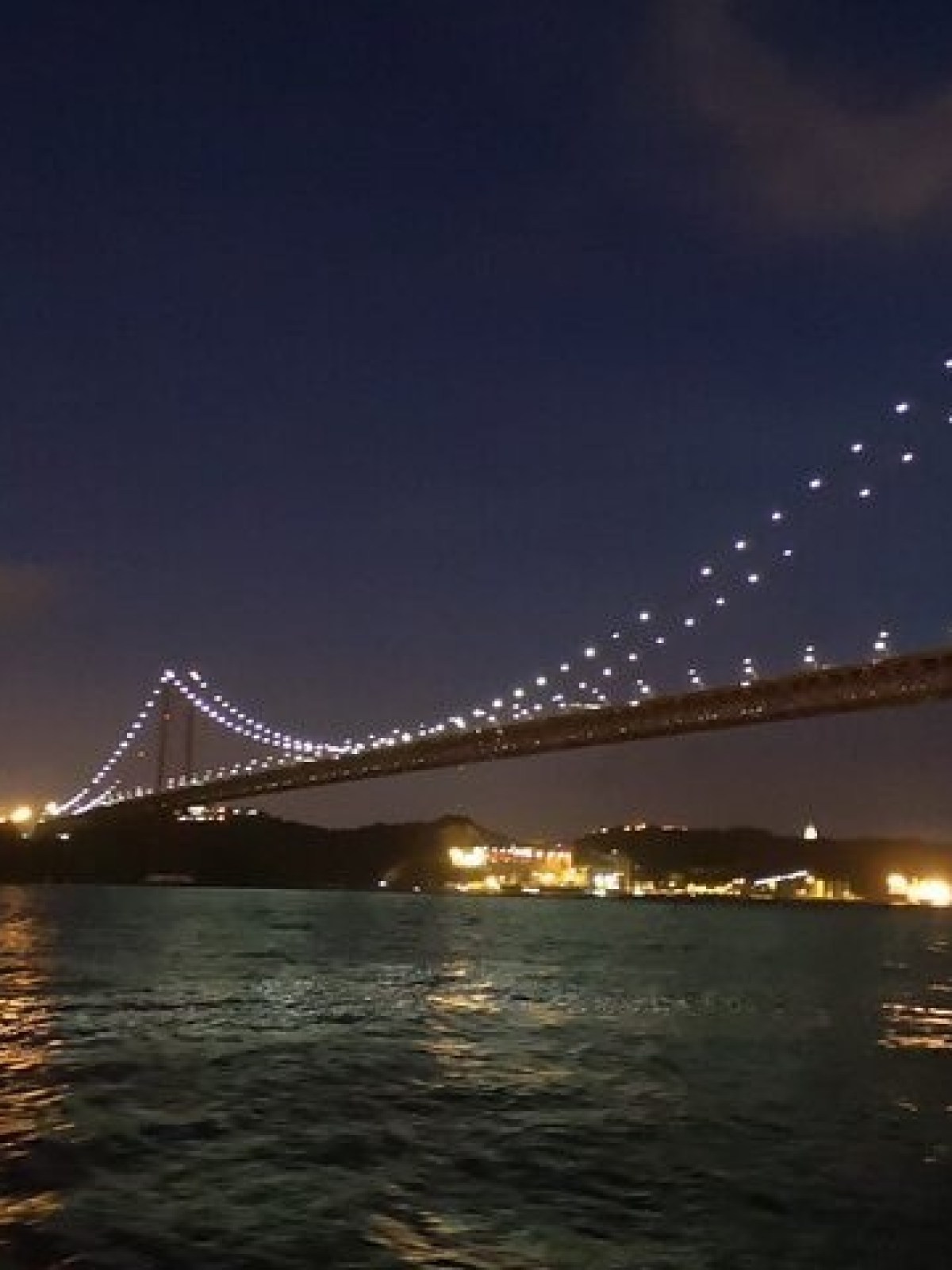 Illuminated suspension bridge and water at night with distant statue.