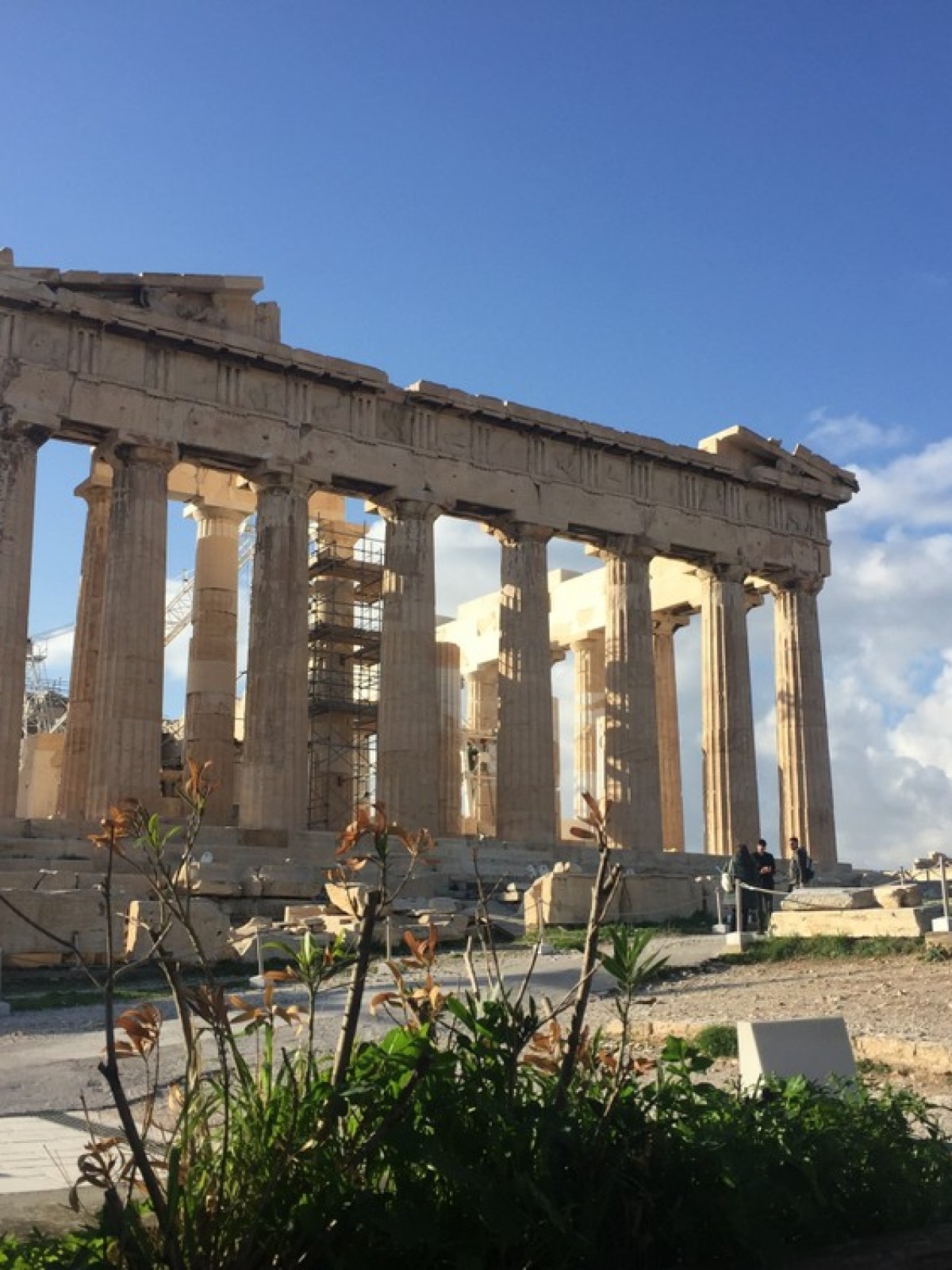 Ancient Greek temple with columns under a sunny sky, surrounded by ruins and some greenery.