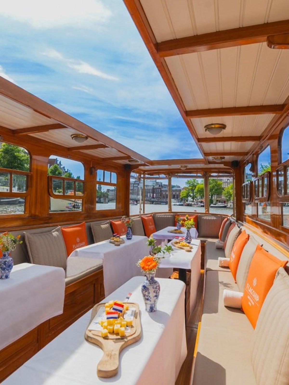 Interior of a wooden boat with tables, flowers, and orange cushions, sailing on a sunny canal.