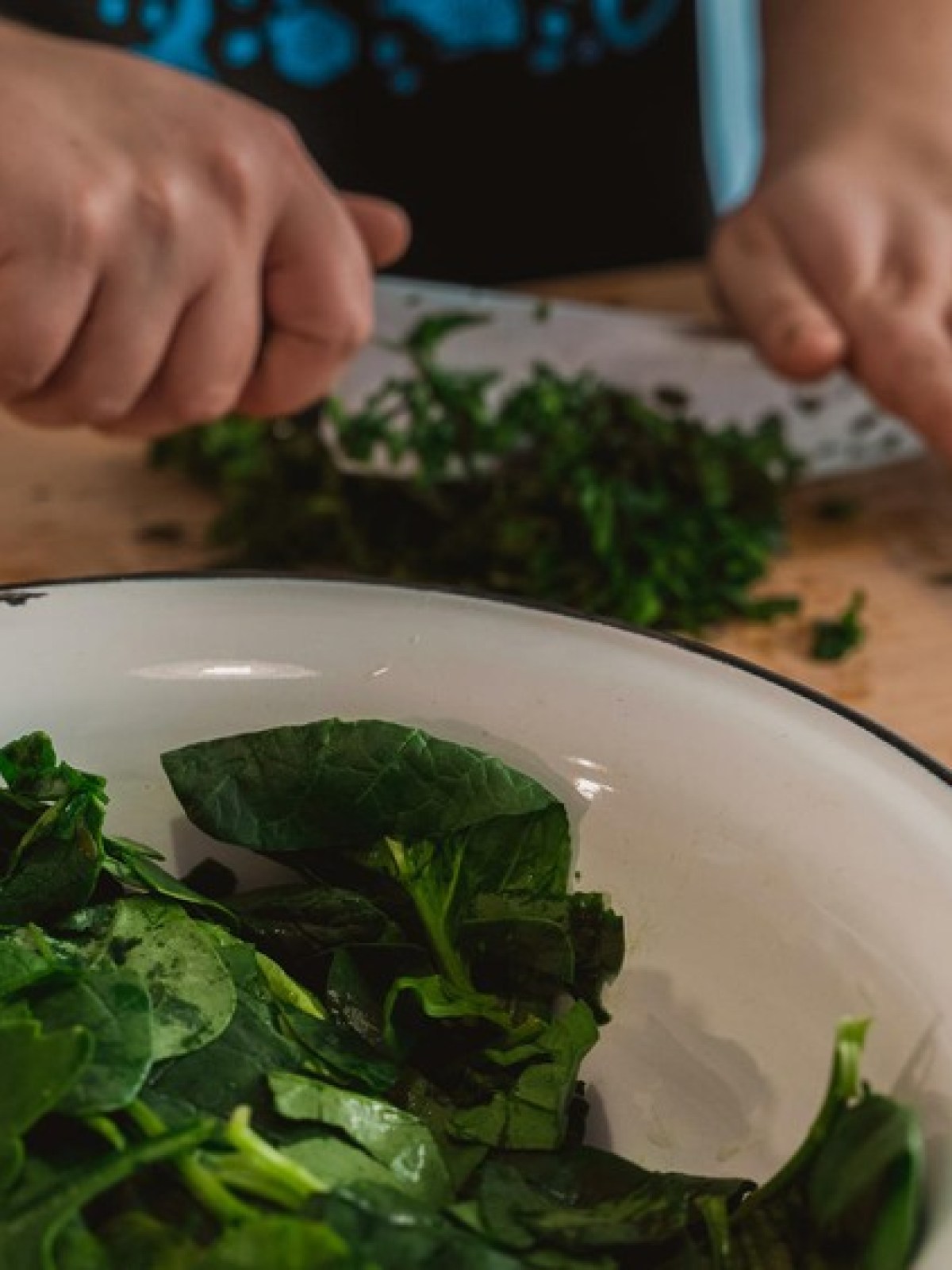 Person chopping spinach on a wooden board with a bowl of leaves nearby.