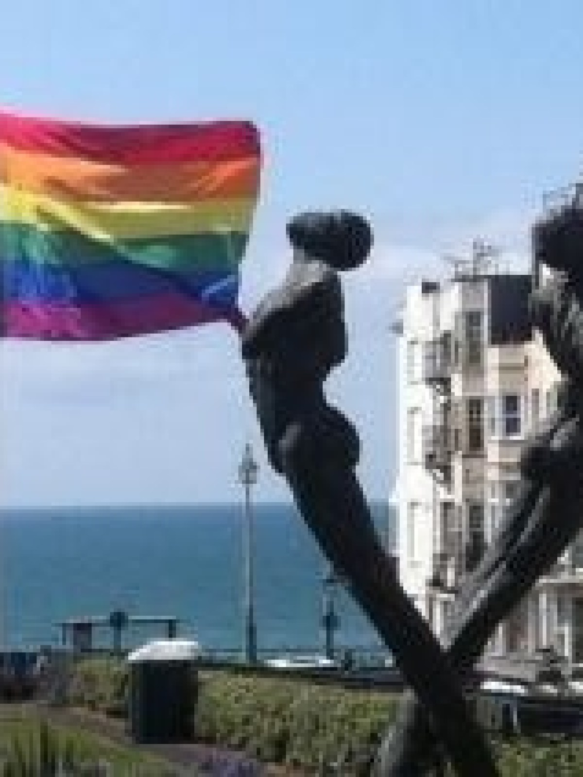 Rainbow flag near two bending statues and seaside buildings on a sunny day.