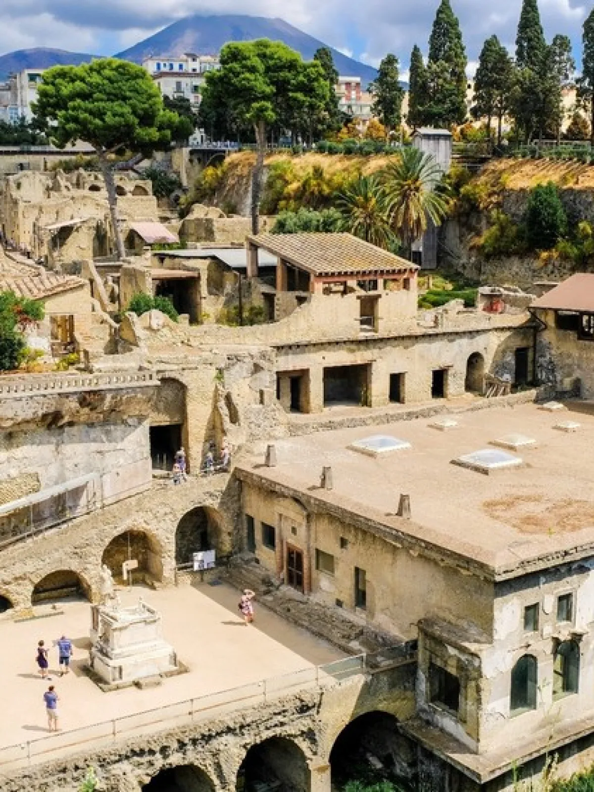 Ancient ruins with stone buildings, trees, and people exploring, set under a cloudy sky.