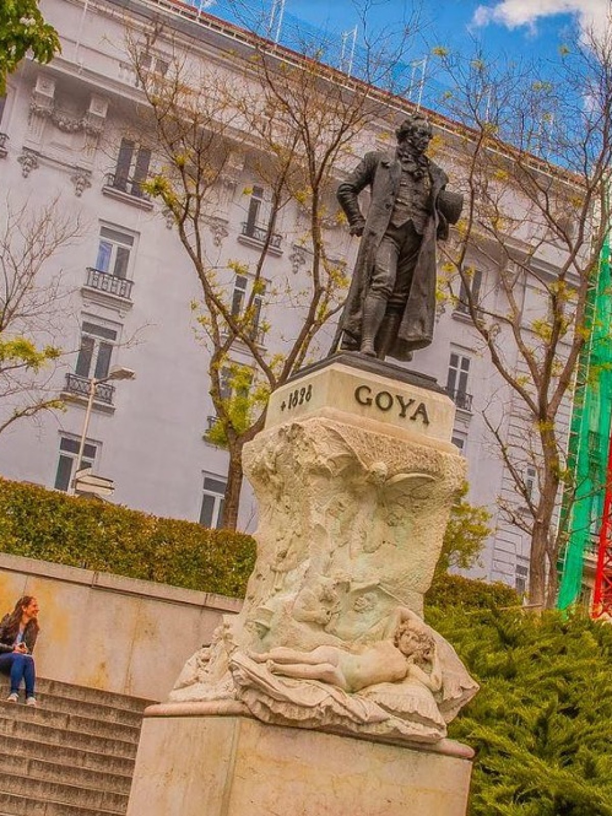 Statue of Goya near stairs, with people sitting, trees, and buildings in the background.