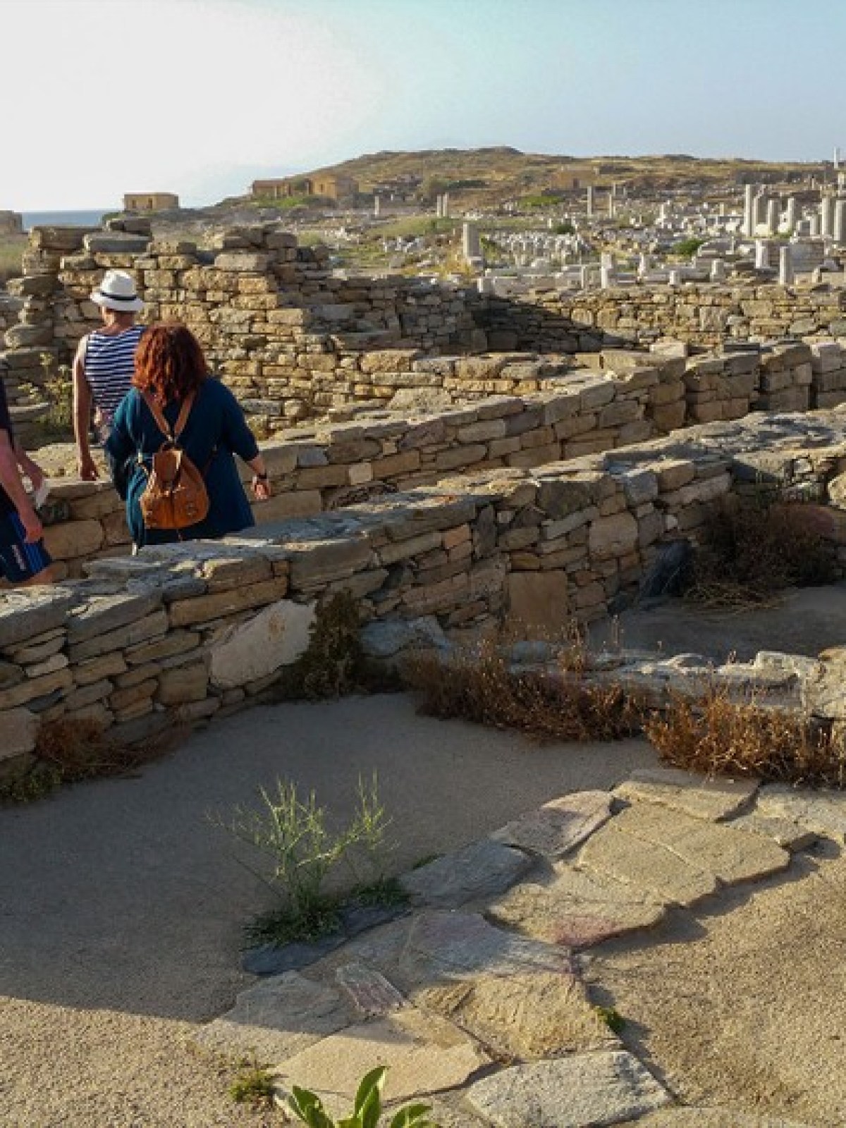 Tourists walk through ancient stone ruins with distant sea view on a sunny day.