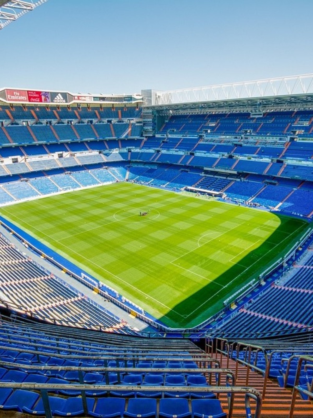Large empty football stadium with blue seats and green pitch under clear sky.