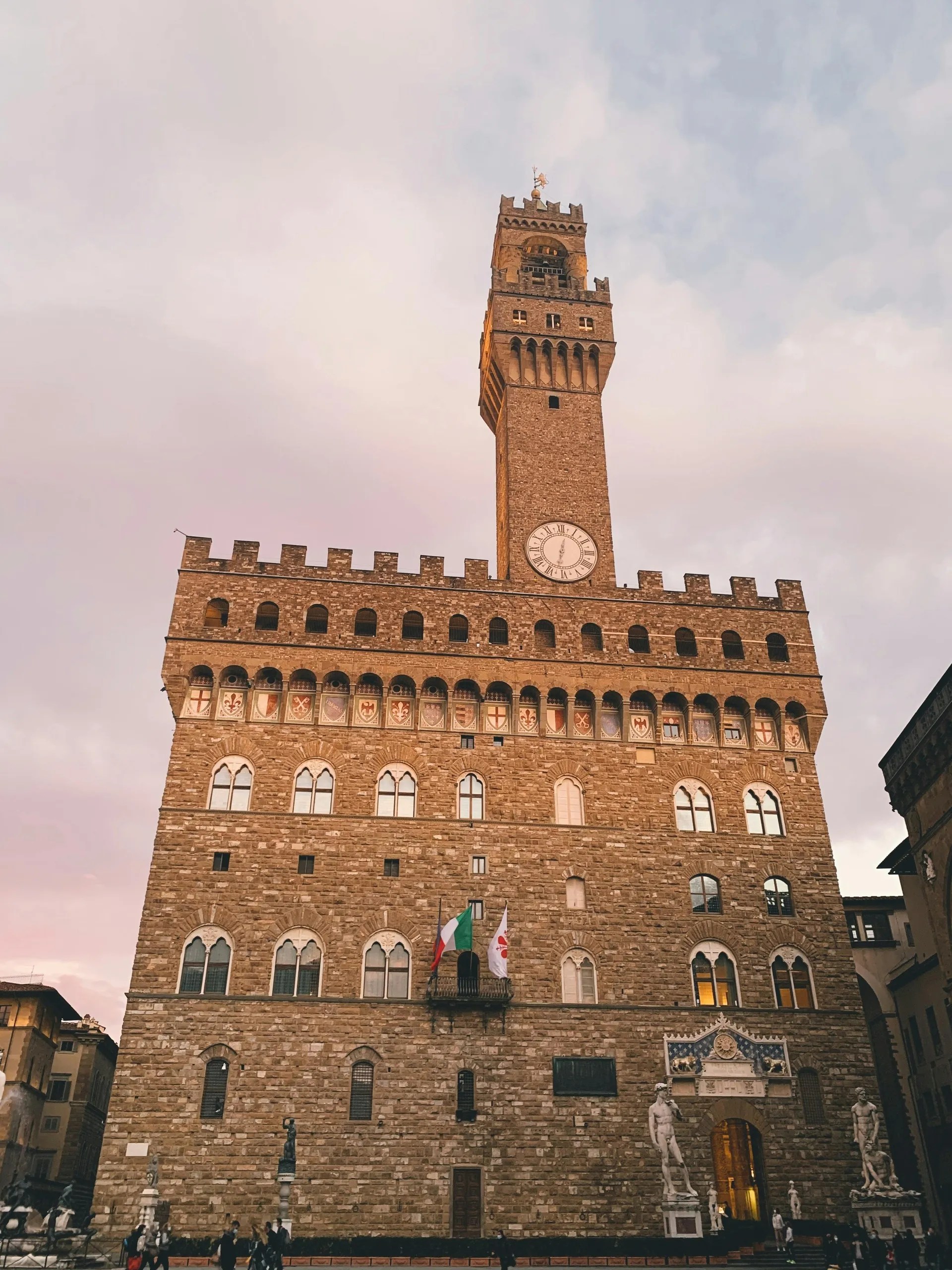 Historic stone building with a tall clock tower in evening light, flags visible.