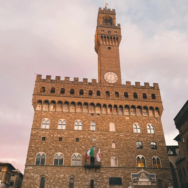 Historic stone building with a tall clock tower in evening light, flags visible.
