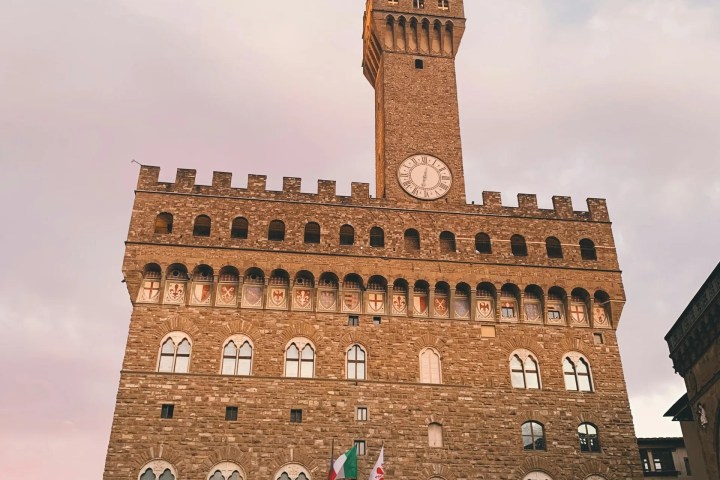 Historic stone building with a tall clock tower in evening light, flags visible.