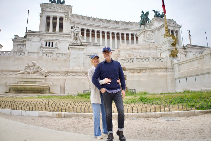 Couple posing in front of a historic building with columns and statues on a cloudy day.