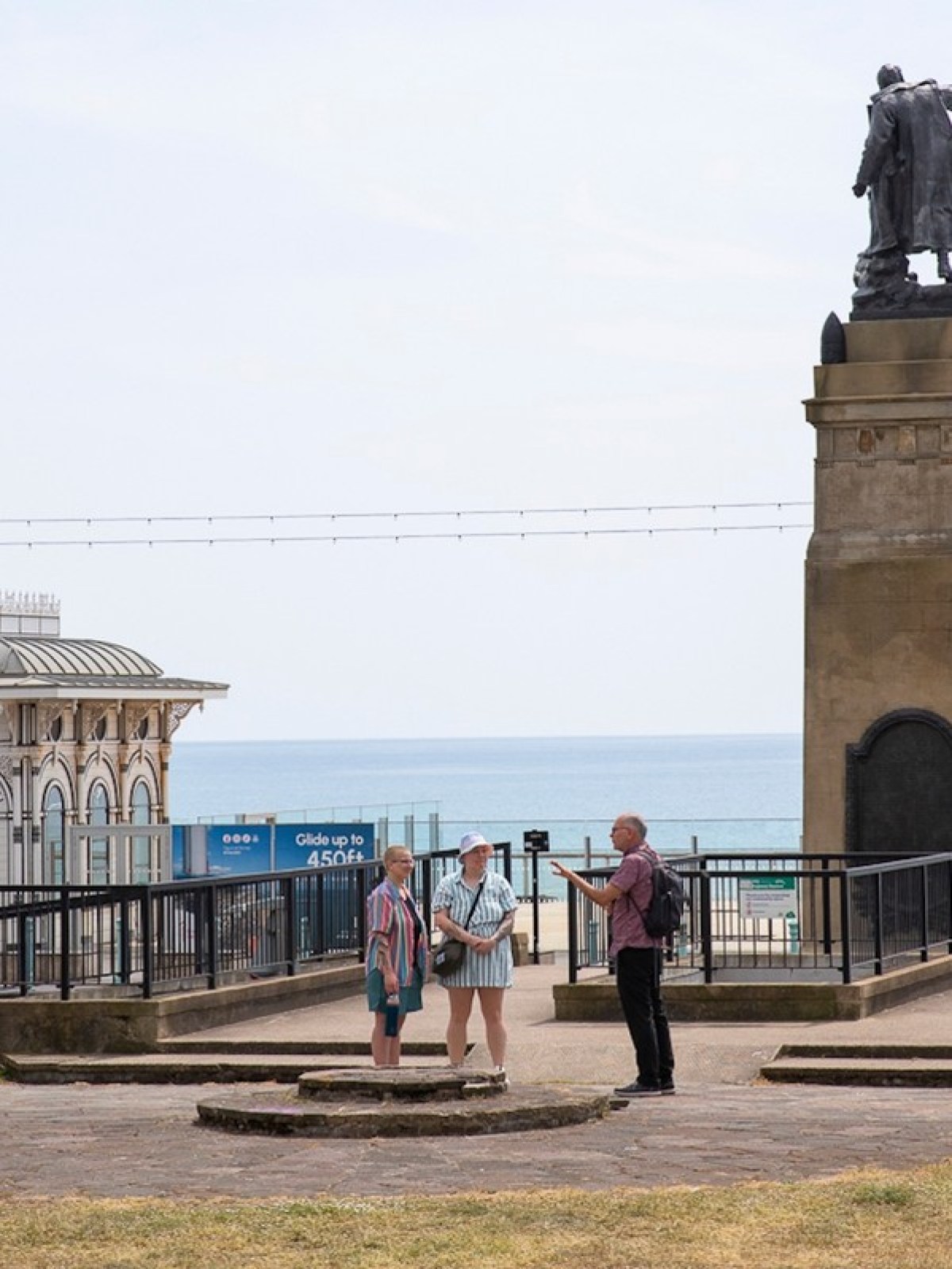 People standing by a seaside statue with ocean and buildings in the background.