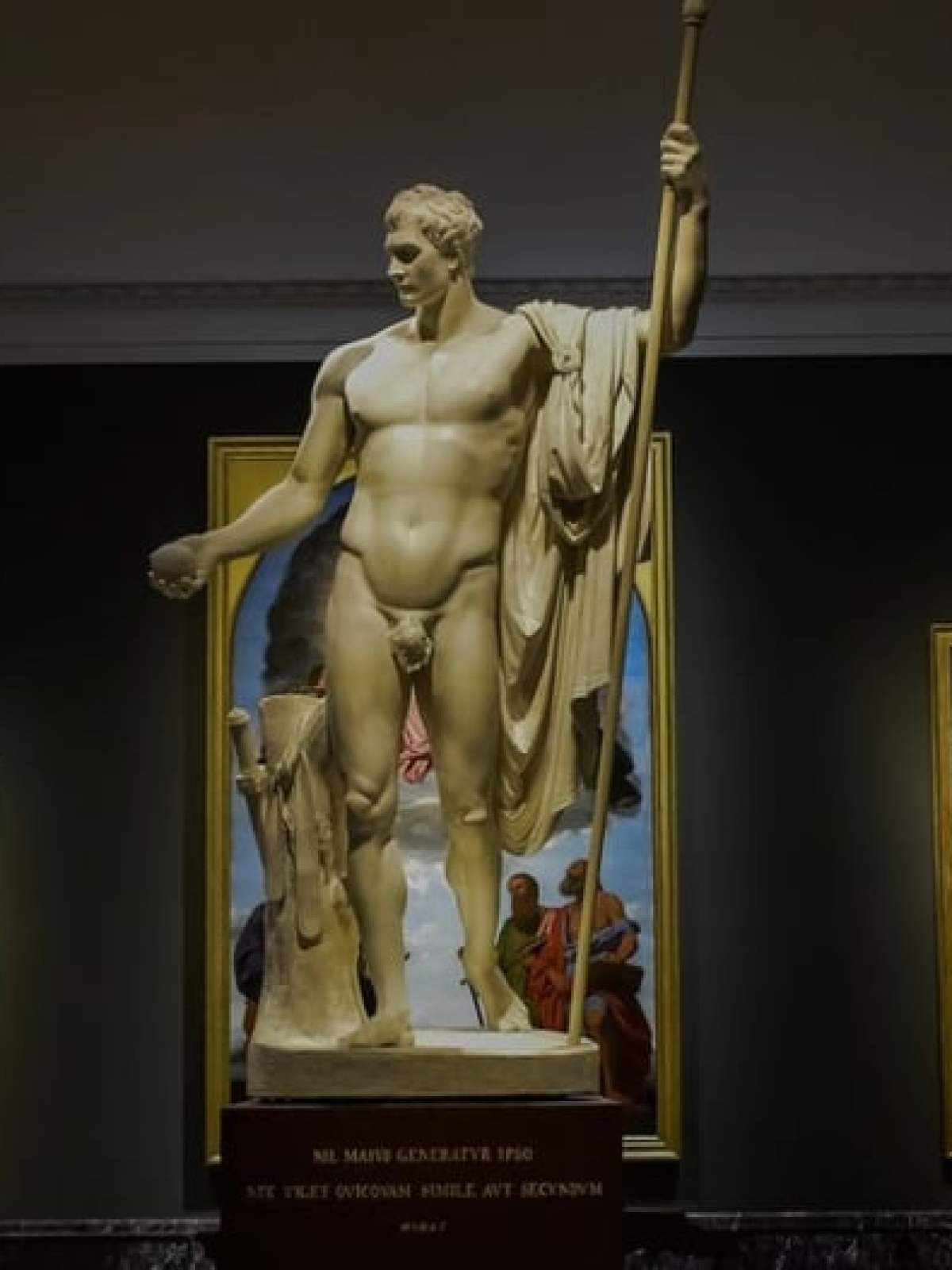 Marble statue in museum surrounded by classical paintings on dark walls.